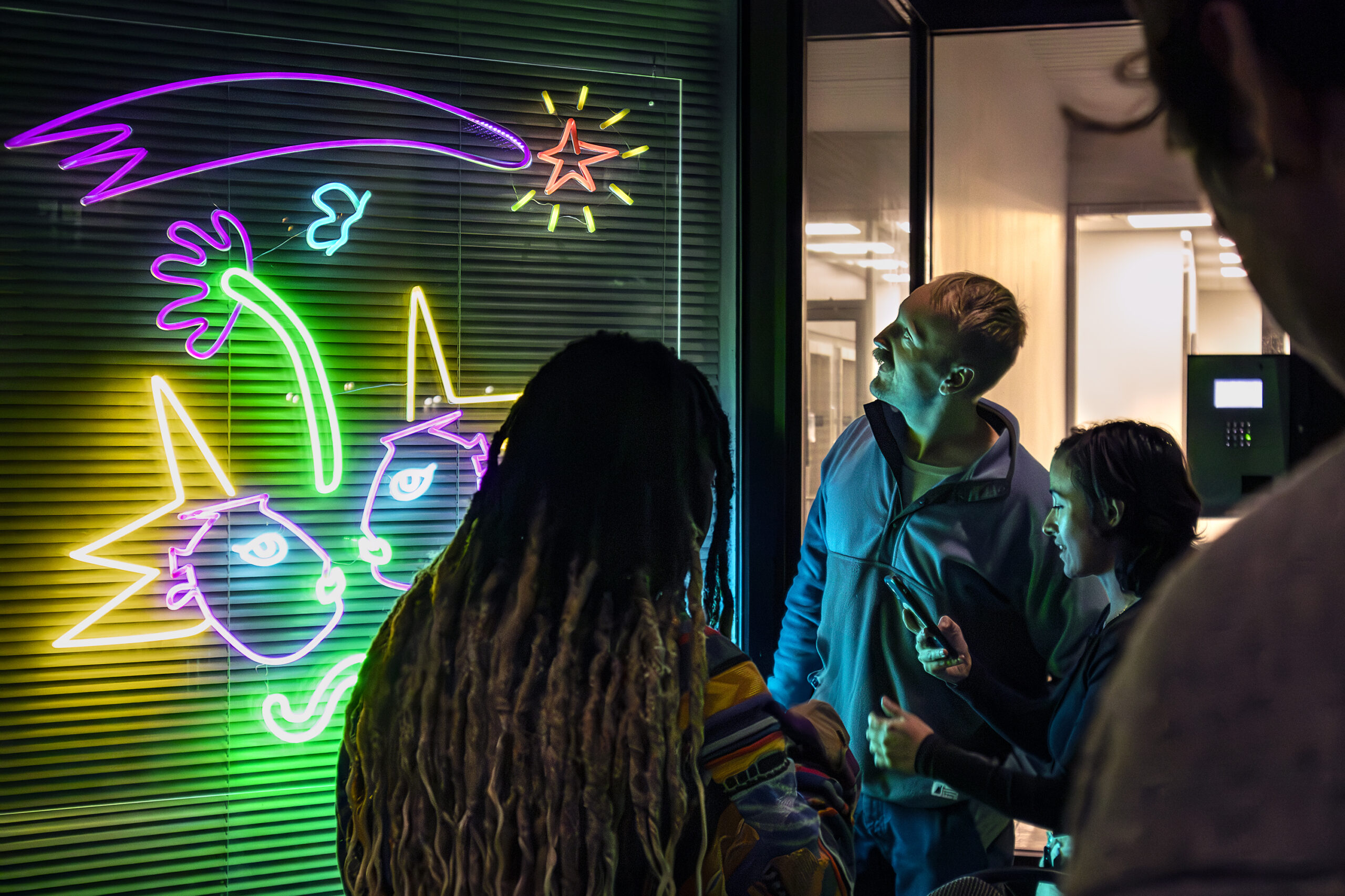 An audience gazing at a public art piece with glowing neon lights in the outline of fun shapes and figures on a window