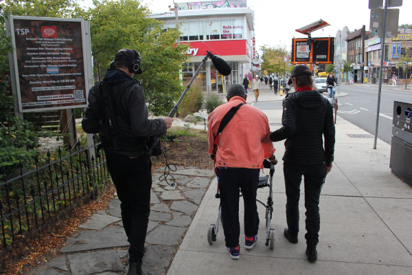 A person guiding another person on a sidewalk who is walking with a walker. A third person is recording audio with a boom mic as part of STEPS Public Art's research on accessibility in art and placemaking