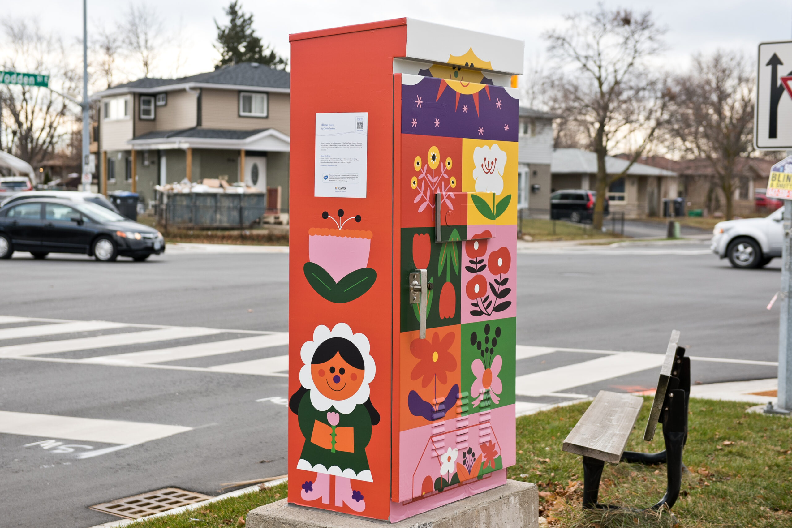 Photo of a traffic box with artwork by Camilla Teodoro; it features a grid/quilt composition with motifs of flowers and wildlife and some characters. It has colours of orange, green, and pink prominently.