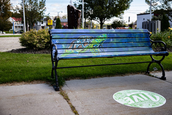 A public bench painted blue with a turtle and a sidewalk decal by artists Ray Vidal and Rhonda Franks for Downtown Tillsonburg and STEPS Public Art
