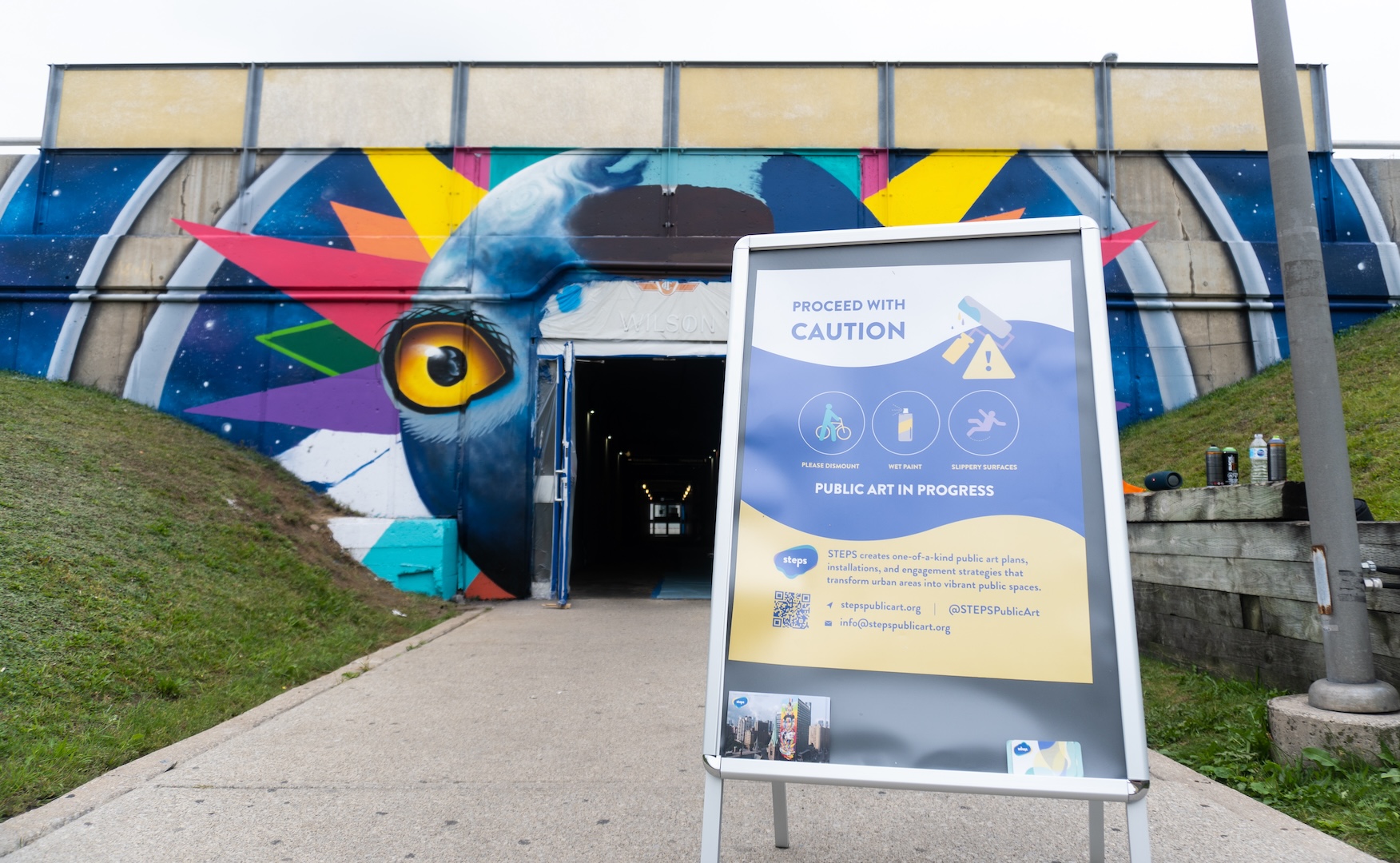 A mural in progress at Wilson TTC Station by artist Shalak Attack. A sandwich board sign informs the community about the project.