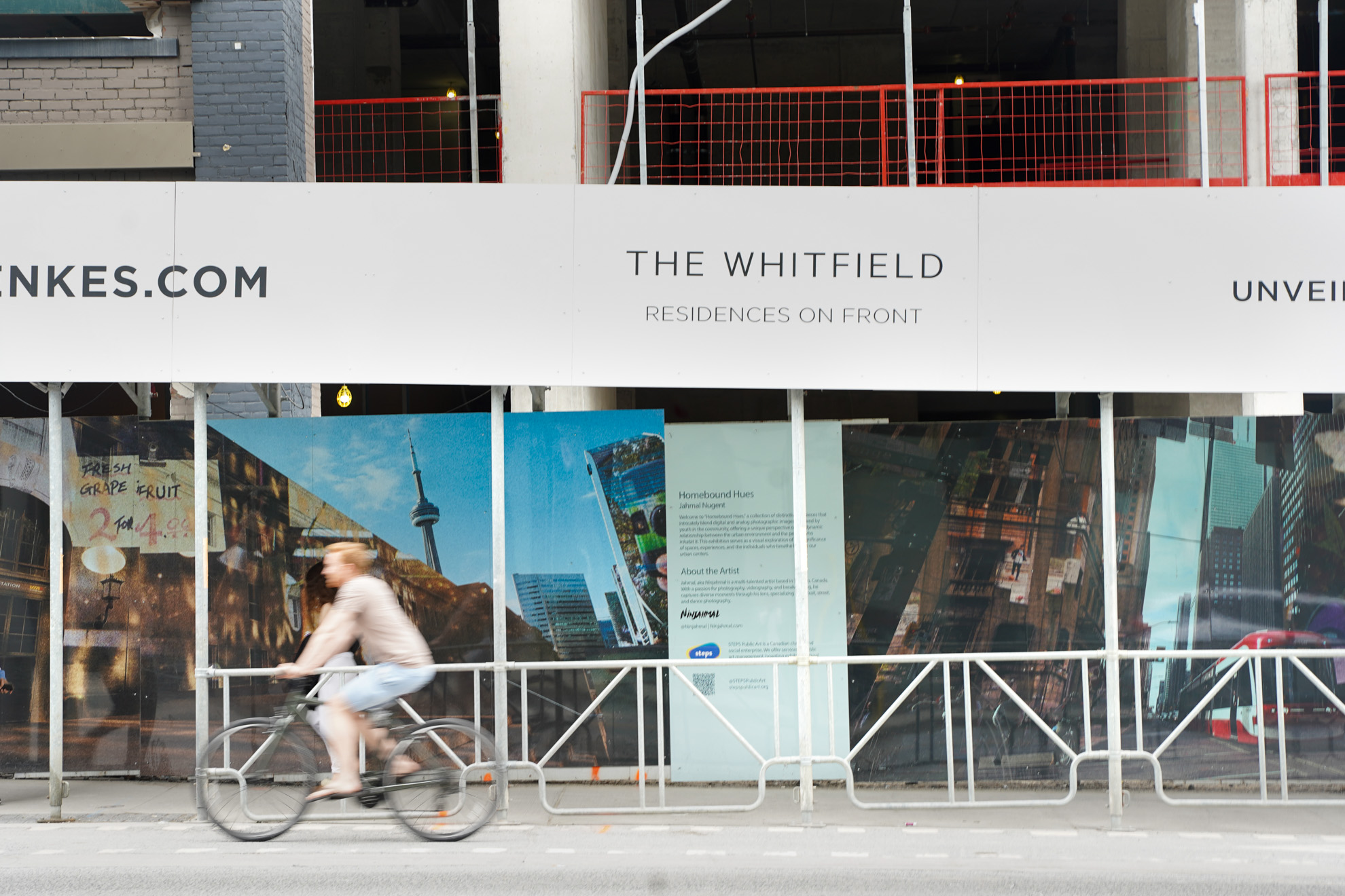 Photo of The Whitfield hoarding artwork, “Homebound Hues” by Jahmal Nugent for STEPS and Menkes Development, from across the street while someone on a bike is cycling passed.