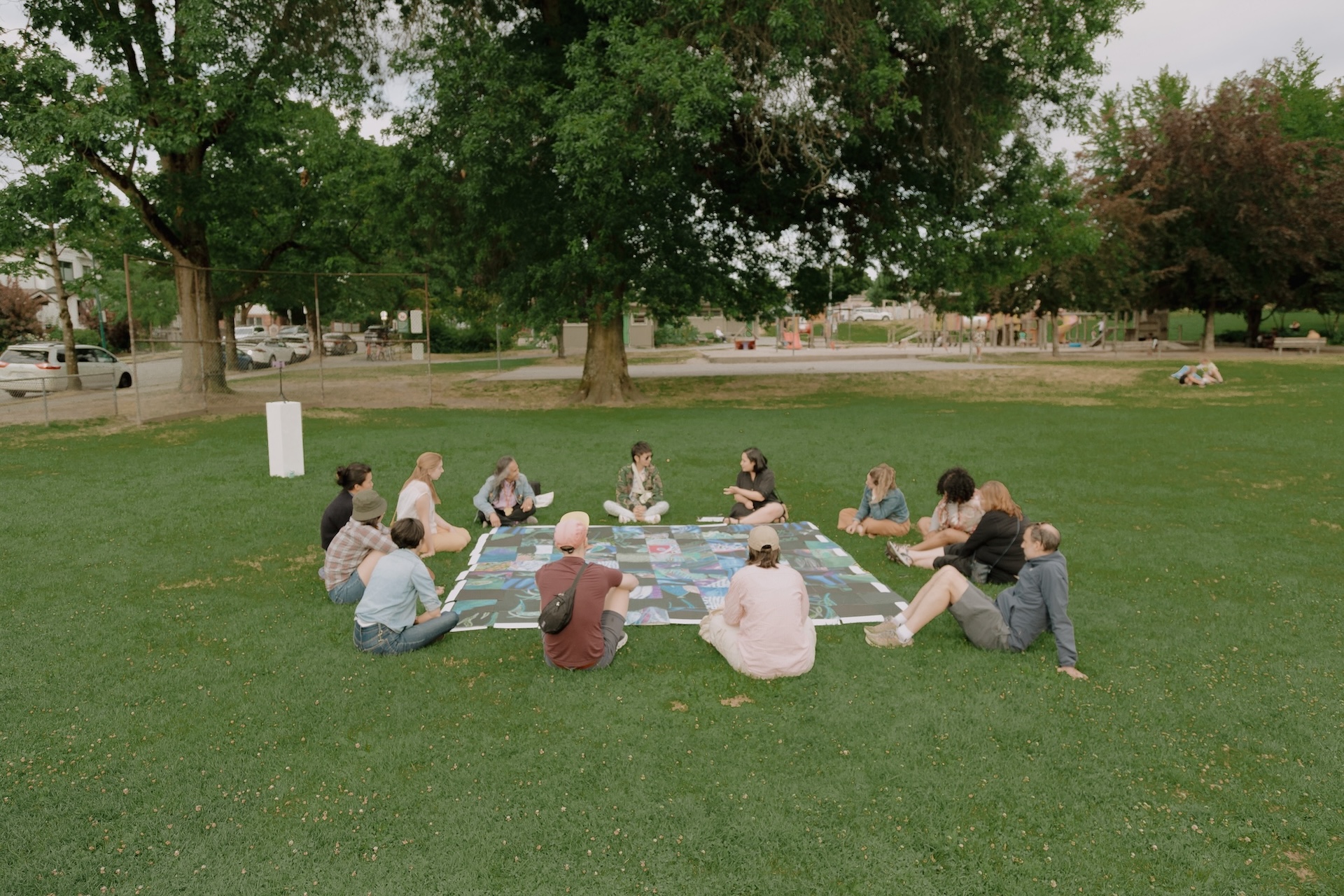 Community members sitting outside in a park and in a circle around a woven placemat as part of Aaniya Asrani's CreateSpace Public Art Residency project