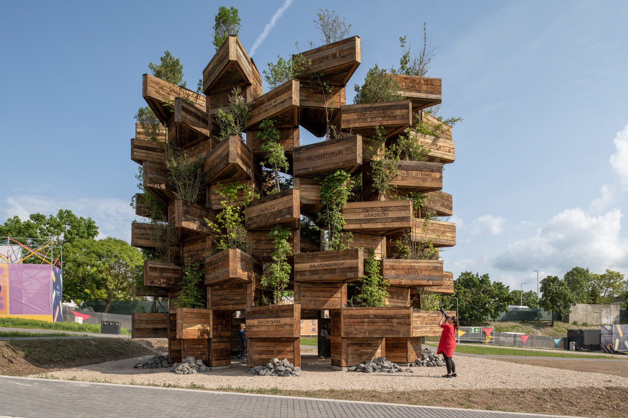 A public art installation of large wooden planters stacked on top of each other with saplings growing. A person is admiring and taking a photo from below.